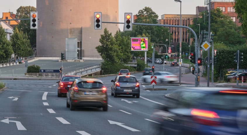 Straße mit Autos an einer Kreuzung bei Grün, im Hintergrund ein großer runder Turm und Ampeln.