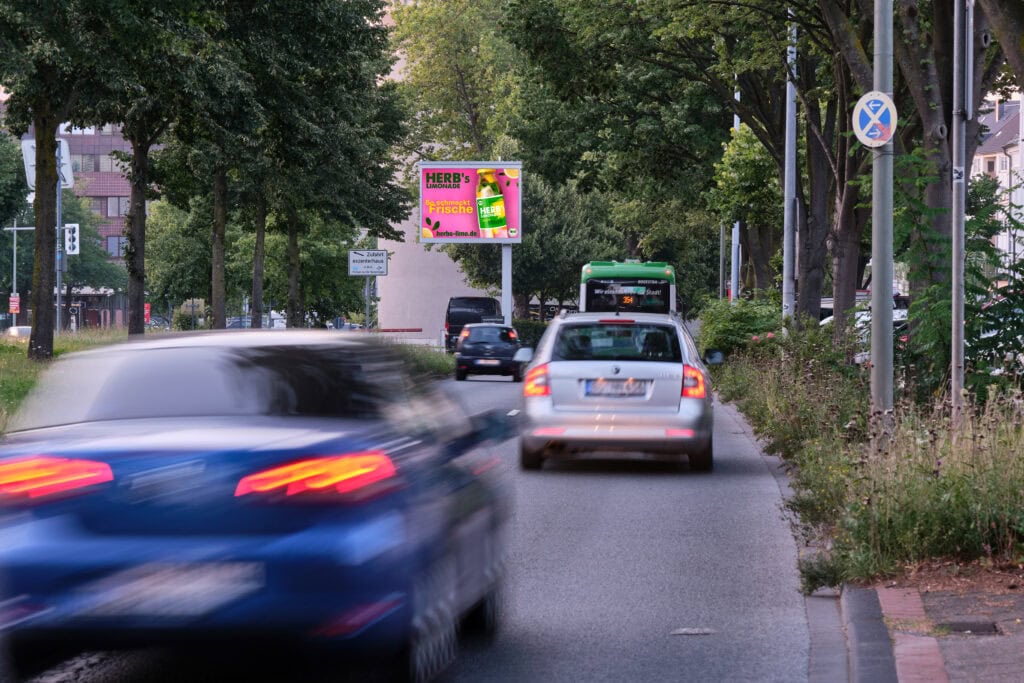 Straße mit Autos, Bäumen und grünem Bus. Plakat mit auffälligen Farben im Hintergrund.