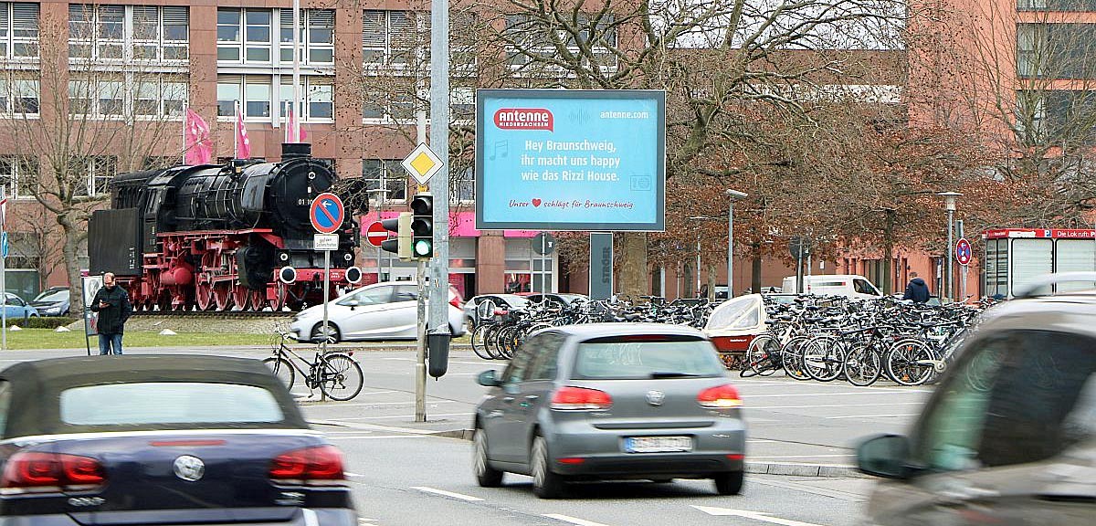 Straßenszene mit Autos, Fahrrädern und einer alten schwarzen Lokomotive im Hintergrund.