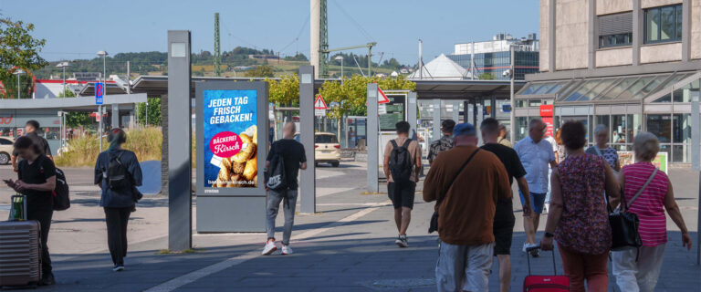 Mehrere Personen auf einem belebten Bahnhofsvorplatz bei sonnigem Wetter. Werbetafel im Hintergrund.