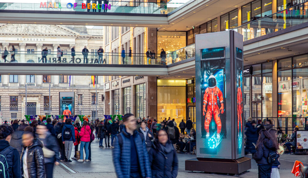 Menschenmenge in der Mall of Berlin. Digitale Displays zeigen einen Astronauten in rotem Anzug.