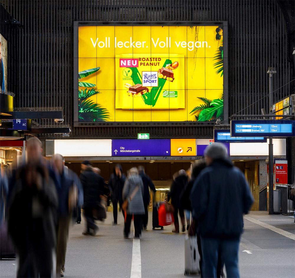 Verkehrsknotenpunkt mit Menschen in Bewegung; großer, bunter Schokoladenwerbung im Hintergrund.