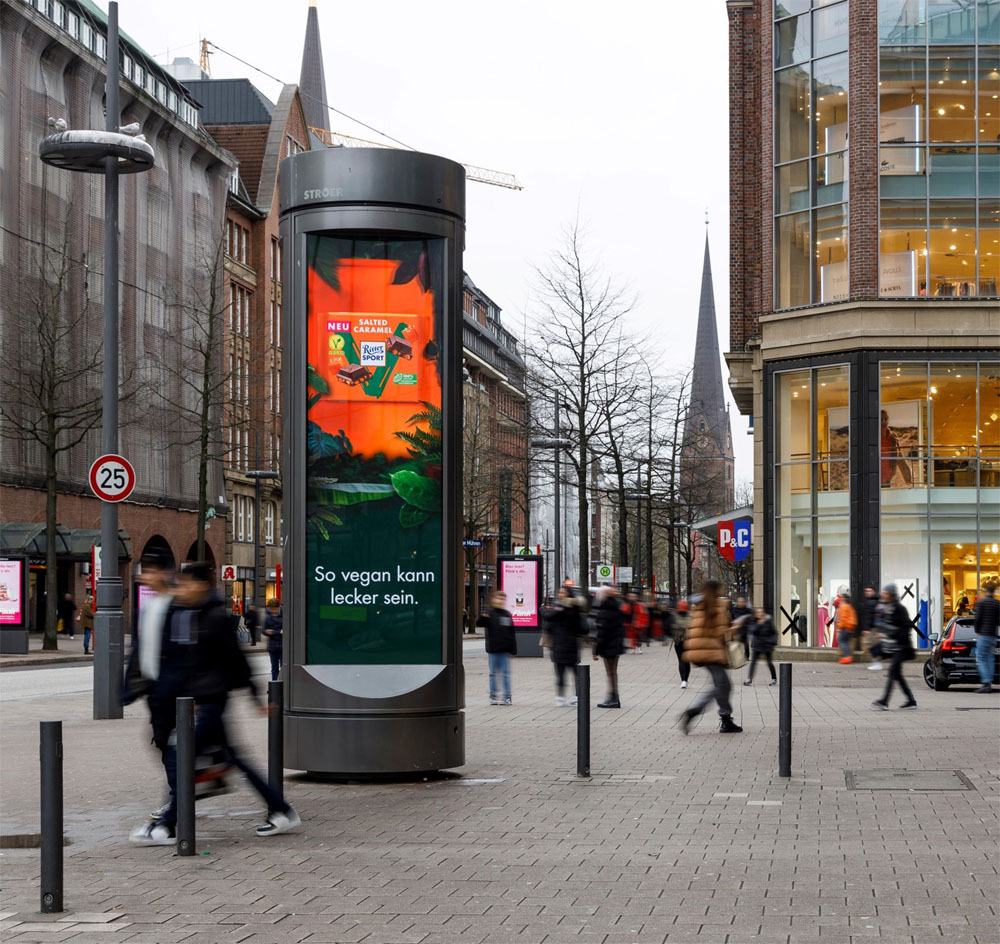 Menschen laufen an einer Werbetafel in einer belebten Straße in einer modernen Stadt vorbei.