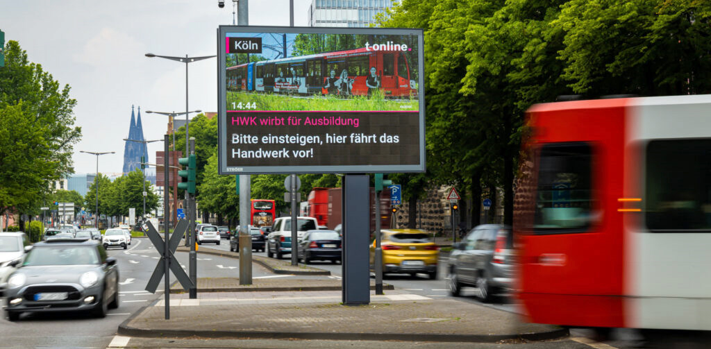 Straßenszene in Köln mit digitalem Werbeplakat für Handwerksausbildung; Sommerwetter, Verkehrsfluss.