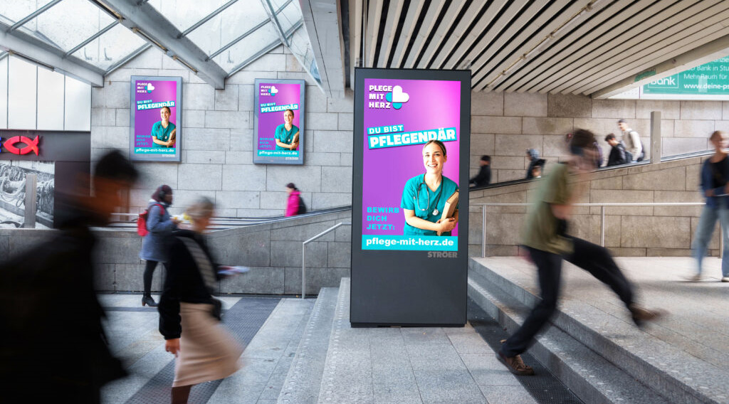 Eine lächelnde Person in Arbeitskleidung auf einem Werbeplakat in einem belebten U-Bahnhof.