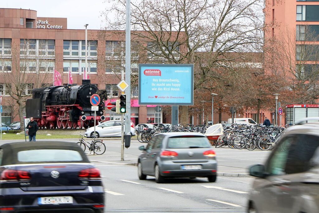 Eine historische Dampflok parkt vor einem Gebäude; im Vordergrund fahren Autos an Fahrrädern vorbei.