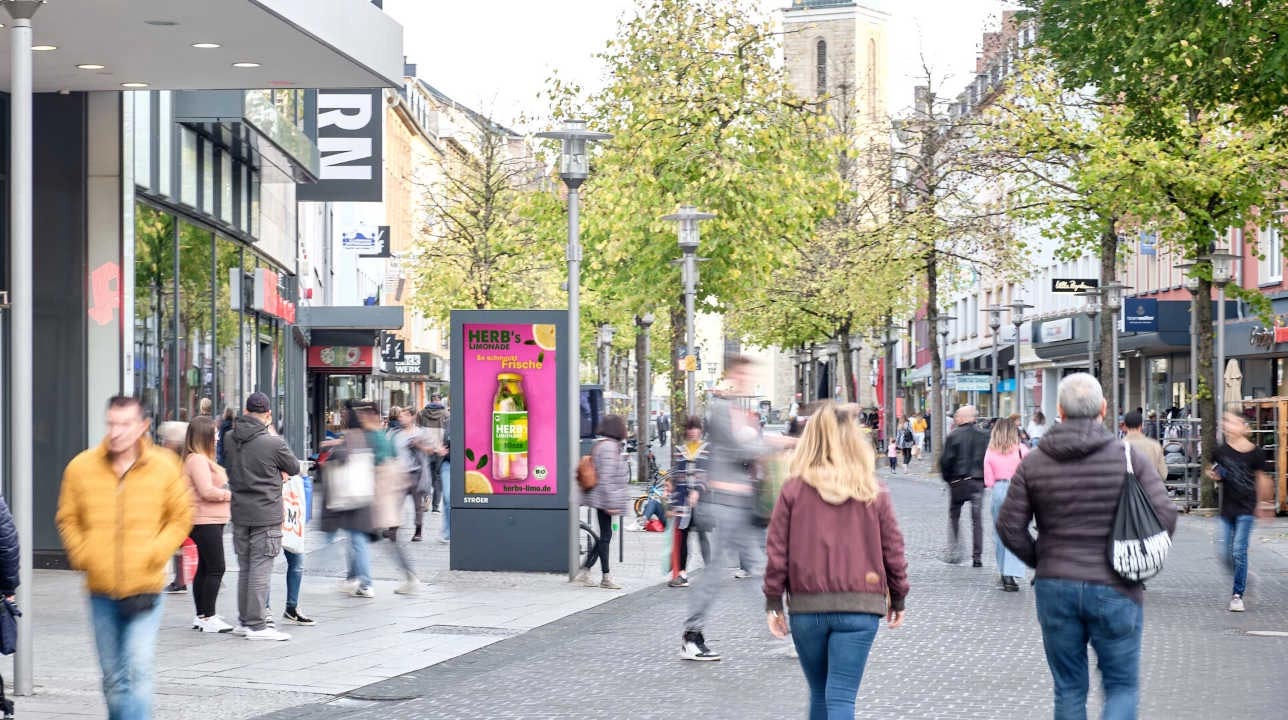 Menschen spazieren durch eine belebte Einkaufsstraße mit Bäumen und Geschäften bei Tageslicht.