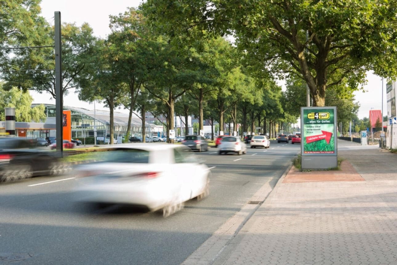 Eine belebte Straße mit fahrenden Autos und Werbetafel, umgeben von Bäumen.