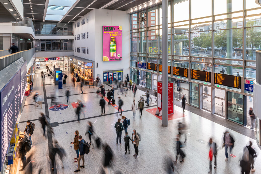 Großer, heller Bahnhof mit Menschen in Bewegung; Werbeplakat und Anzeigetafeln sichtbar.